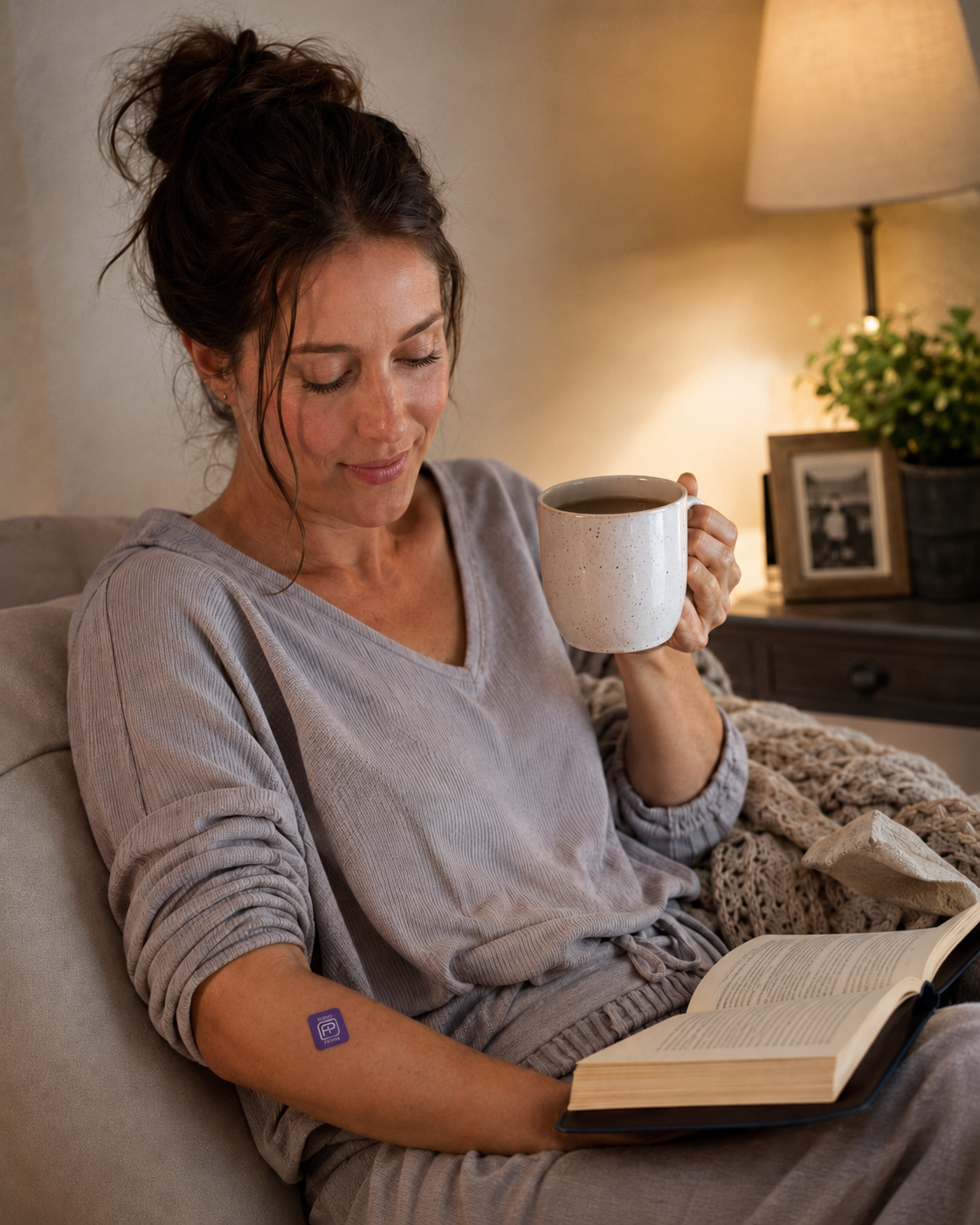 Woman on a sofa at night holding a mug, wearing a purple Bedtime patch on her forearm